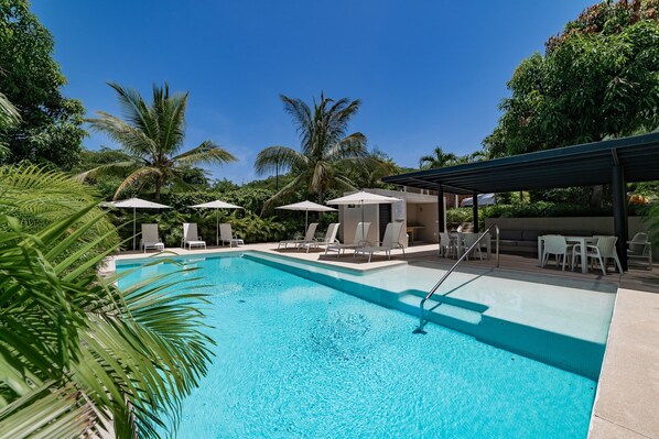 A swimming pool with lounge chairs and umbrellas, surrounded by palm trees and greenery.