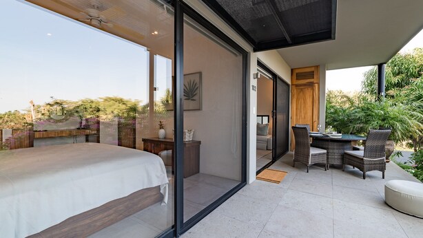 Une chambre moderne dotée d’un grand lit, d’un mur en verre et d’un balcon aménagé avec du mobilier d’extérieur.