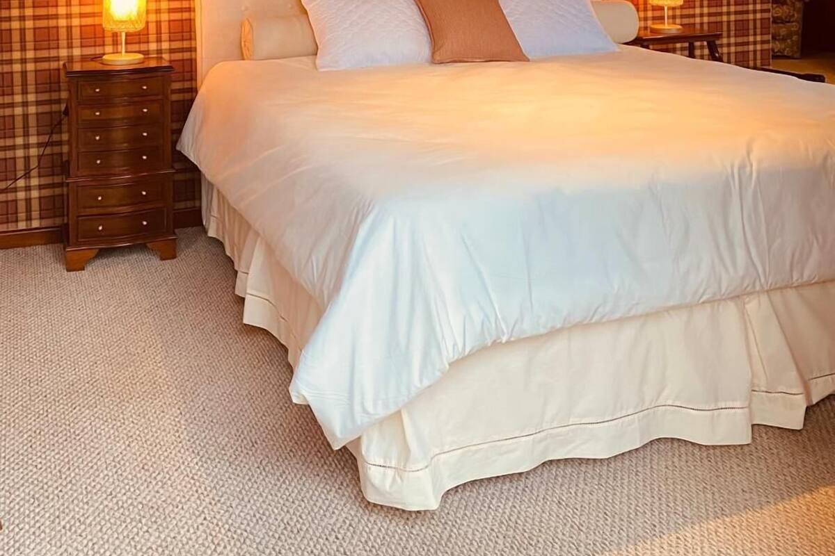 A bedroom with a plaid-patterned wall, a large bed with white bedding, two bedside lamps, and a wooden ceiling.