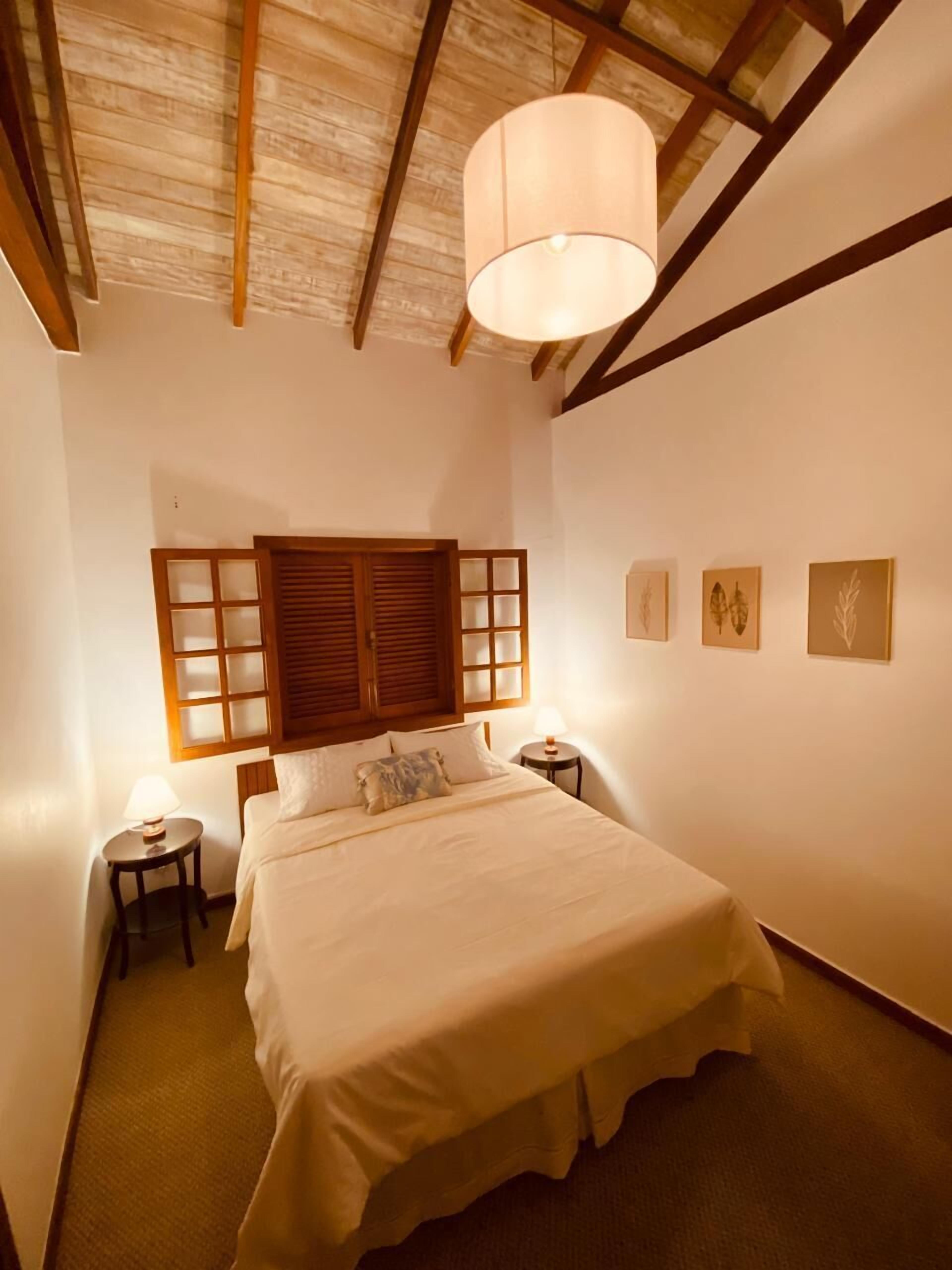 Um quarto com cama, teto de madeira, janela com persianas e luminária fixada na parede.