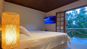 Ein Schlafzimmer mit Bett, Nachttisch mit Lampe, Fernseher an der Wand und Blick auf Bäume durch eine Glastür.