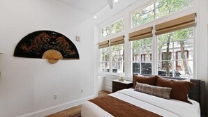 Une chambre à coucher avec un lit, un ventilateur décoratif fixé au mur et de grandes fenêtres donnant sur l’extérieur.