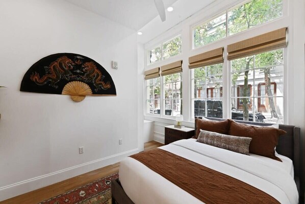 A bedroom with a bed, a wall-mounted decorative fan, and large windows with a view of the outside.