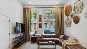 Un salon avec un canapé en cuir marron, une table basse, un téléviseur posé sur un meuble noir et une grande fenêtre avec des rideaux.