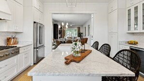 A modern kitchen with white cabinetry, a central island, and a dining area with wicker chairs. - 7BR Coastal Retreat | Heated Pool & Game Area (Bay St. Louis)