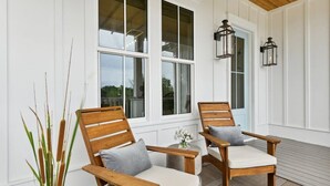 A covered outdoor seating area with two wooden chairs and a small table, white walls, and large windows. - 7BR Coastal Retreat | Heated Pool & Game Area (Bay St. Louis)
