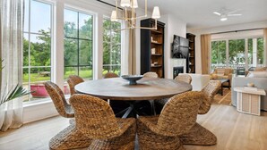 A spacious living room with a round wooden table surrounded by woven chairs, a modern chandelier, and large windows offering a view of the outdoors. - 7BR Coastal Retreat | Heated Pool & Game Area (Bay St. Louis)