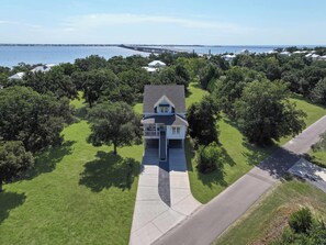 A two-story house with a front porch, surrounded by a well-maintained lawn and trees, with a view of the water and a pier in the background. - 7BR Coastal Retreat | Heated Pool & Game Area (Bay St. Louis)