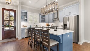 A modern kitchen with a central island, wooden chairs, and a stainless steel refrigerator. - Brand New Home, Sleeps 10, Pool and Screened-in Porch with TV! (St Simons Island)