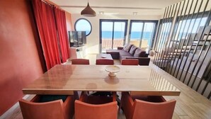 A modern dining area with a wooden table and orange chairs, a red curtain, and a view of the sea through large windows.