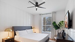Une chambre moderne avec un grand lit, un ventilateur de plafond, une télévision et une fenêtre avec des rideaux.