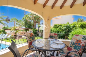 Un área de comedor al aire libre techada con vista a la piscina, que incluye una mesa redonda preparada para cuatro personas, rodeada de sillas de colores.