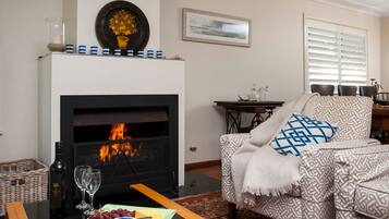 A living room with a fireplace, a sofa with patterned cushions, a small table with a bottle and glasses, and a wall with a framed picture.