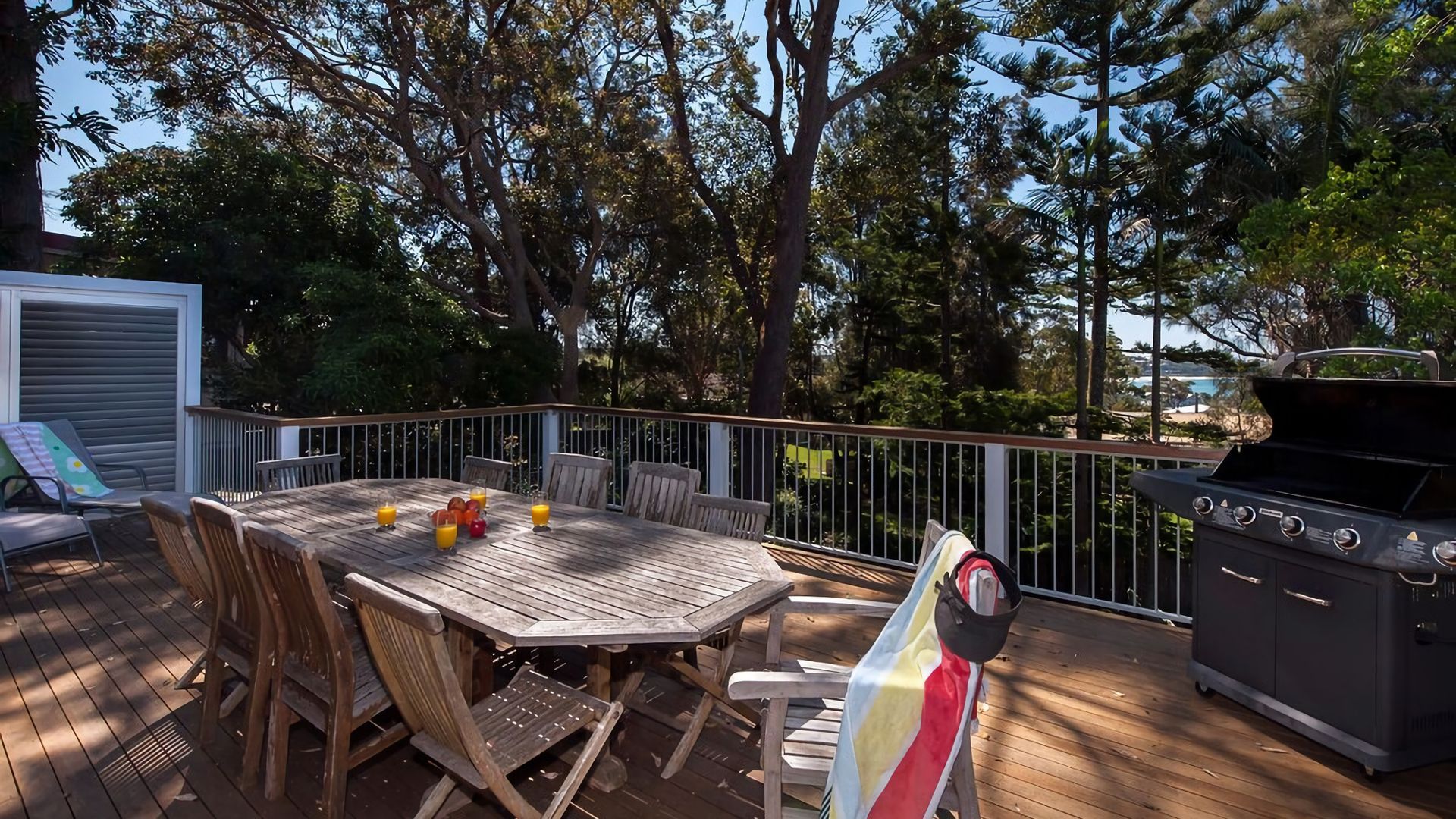 An outdoor deck with a wooden table and chairs, a barbecue grill, and a view of trees and a building in the background.