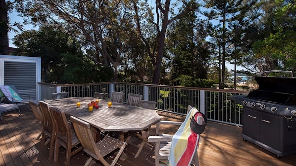 An outdoor deck with a wooden table and chairs, a barbecue grill, and a view of trees and a building in the background. - Vitamin Sea is pet friendly (Mollymook)