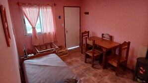 Een kamer met een bed, een houten tafel met stoelen, een klein fornuis en een raam met gordijnen.