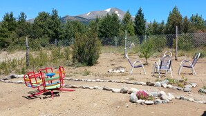 Een speeltuin met een klimconstructie en twee blauwe stoelen in een omheind buitengebied, met een berg op de achtergrond.