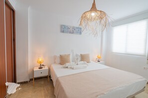 A hotel room with a bed, two bedside tables, a wall-mounted artwork, a hanging light fixture, and a window with blinds.