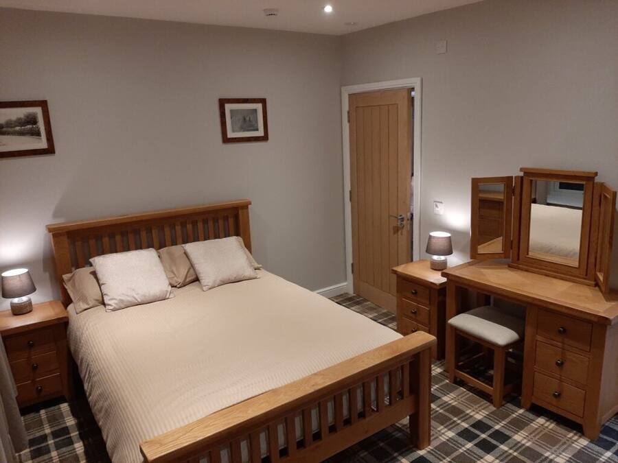 Quarto com cama de madeira, mesa de cabeceira com luminária, cômoda com espelho e uma porta.
