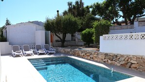 Una piscina rettangolare con acqua cristallina e blu, circondata da pavimento in piastrelle bianche e un muro in pietra. Diverse poltrone da relax bianche sono disposte lungo il bordo della piscina.
