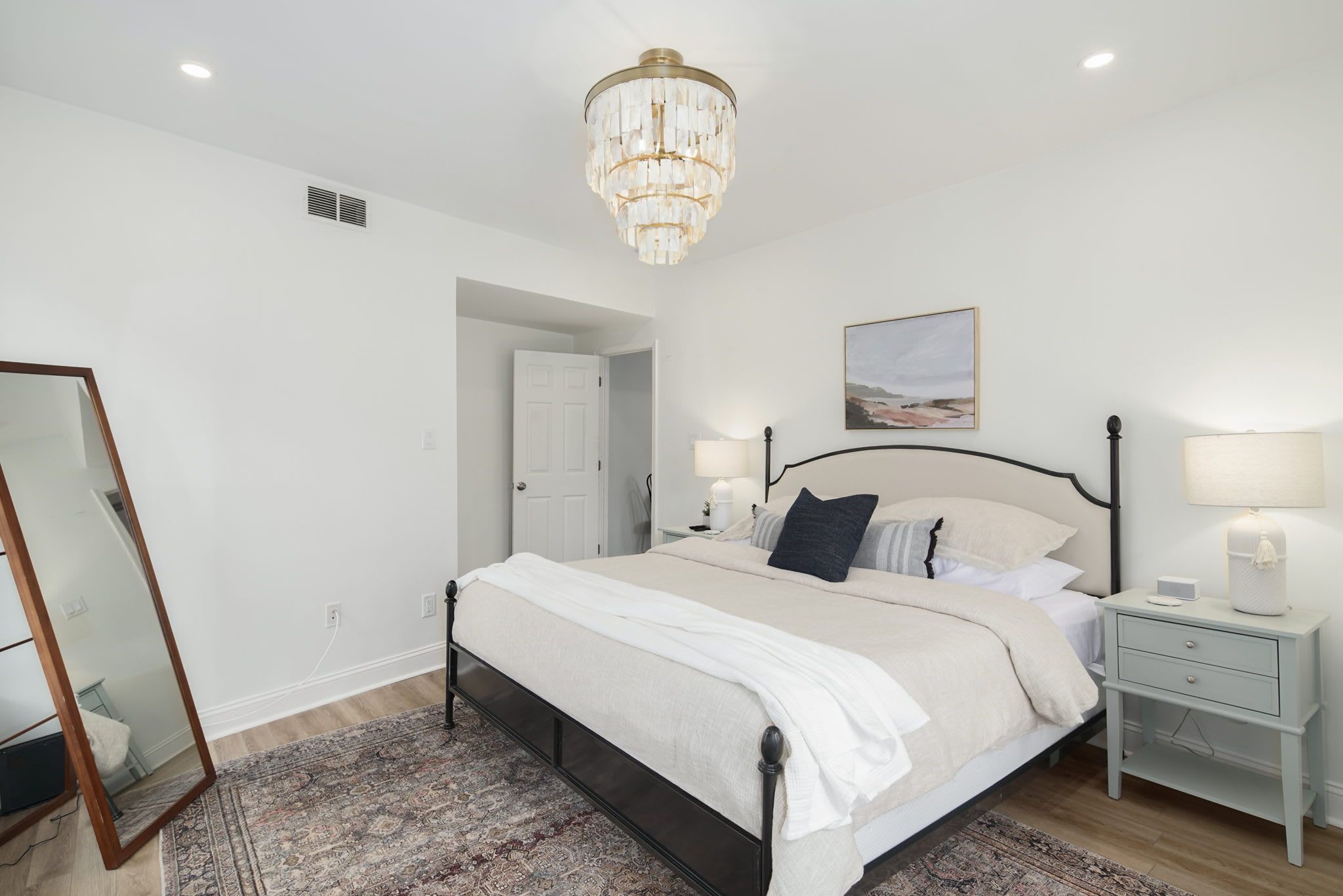 A bedroom with a bed, bedside tables, a chandelier, a mirror, and a door.