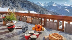 Ein Tisch mit Obstschale, Tassen, Teekanne und Gebäck auf einem Balkon mit Bergblick.