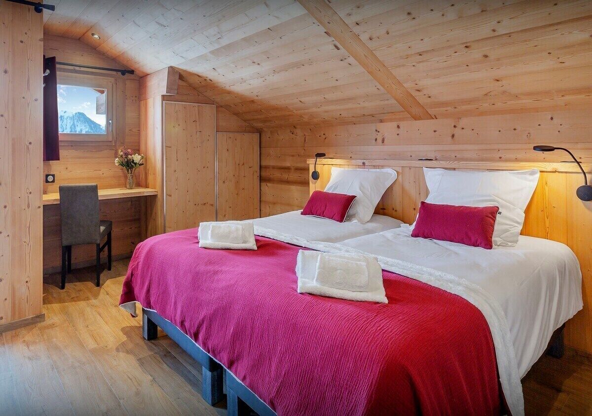 A wooden cabin bedroom with two beds, a desk, and a window with a mountain view.