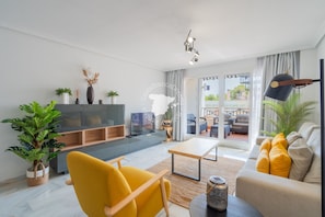 A modern living room with a sofa, armchair, and coffee table. There is a TV on a stand, a plant, and a window with curtains.