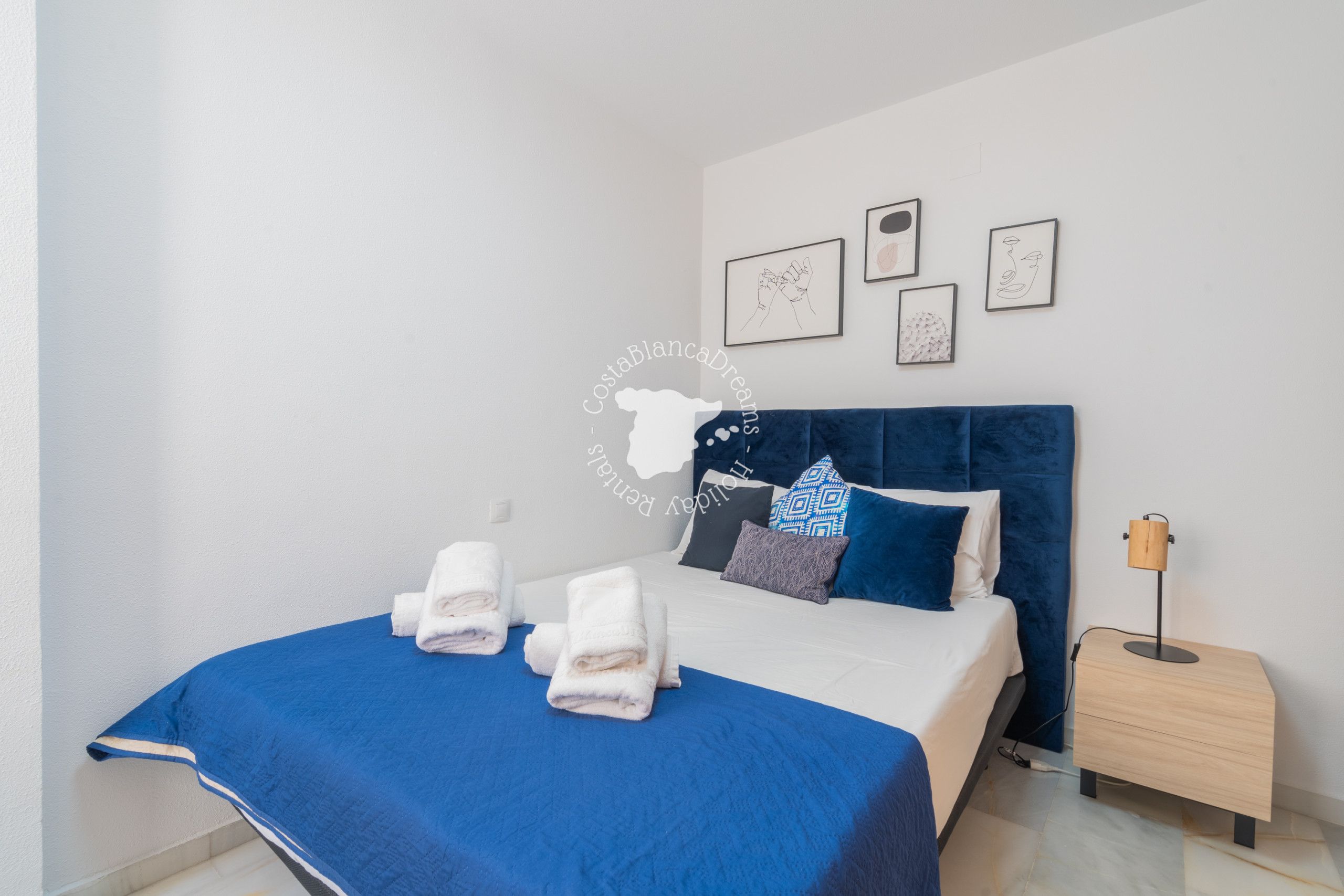 A neatly made bed with blue bedding and white towels, a wooden nightstand with a lamp, and framed artwork on the wall.