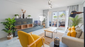 Uma sala de estar moderna com sofá, poltrona e mesa de centro. Há uma televisão em um móvel, uma planta e uma janela com cortinas.