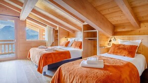 A wooden cabin with two beds, each with orange bedding and white towels, and a balcony with a mountain view.