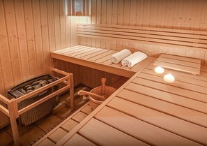 Una sauna de madera con bancos, un banco con toallas, un calentador de piedra y un cubo de madera.