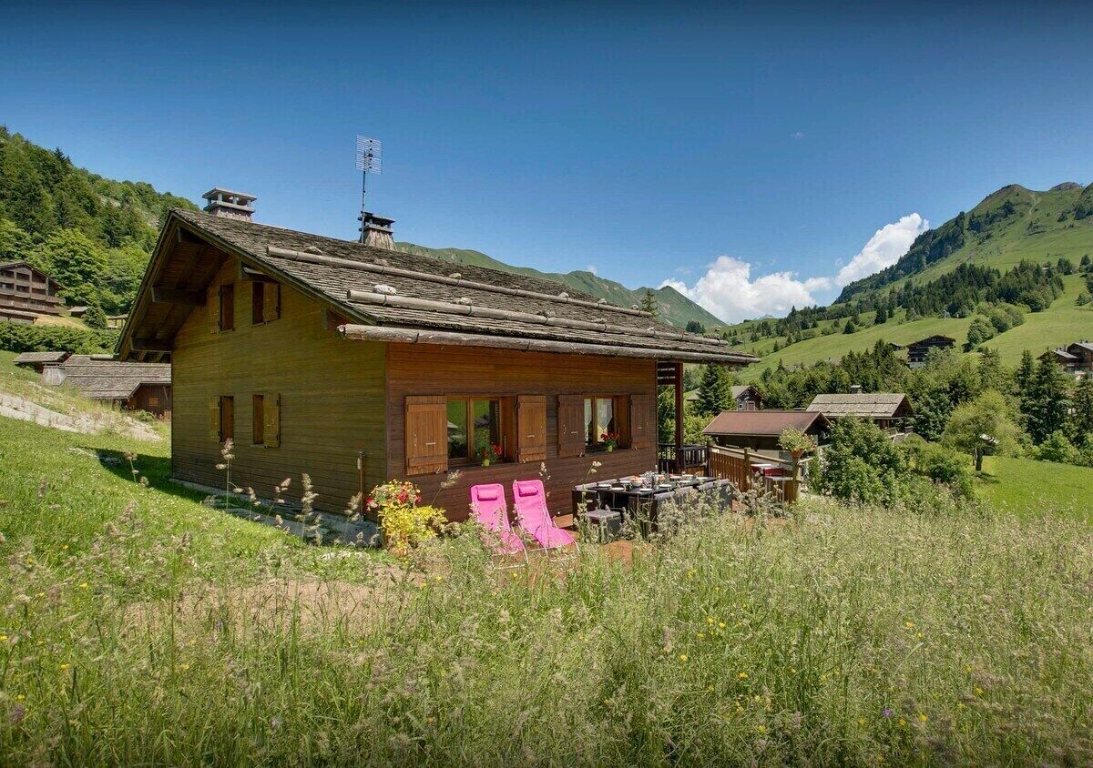 Een houten hutje met terras, omgeven door een grasveld en bergen op de achtergrond.