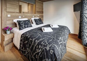 A modern hotel room with a wooden headboard, a large bed with patterned bedding, a small bedside table with a vase of flowers, and a flat-screen TV mounted on the wall. - Jacuzzi hammam and sunny terrace (Le Grand-Bornand)