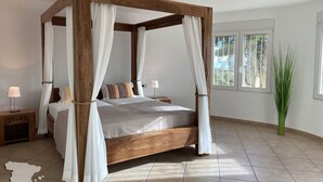 A four-poster bed with white drapes, a bedside table with a lamp, and a large window with a view of trees.