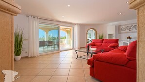 A spacious living room with a red sofa, a glass door leading to an outdoor area, and a kitchen in the background.
