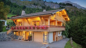A two-story wooden chalet with a balcony, stone foundation, and a view of a mountainous landscape.