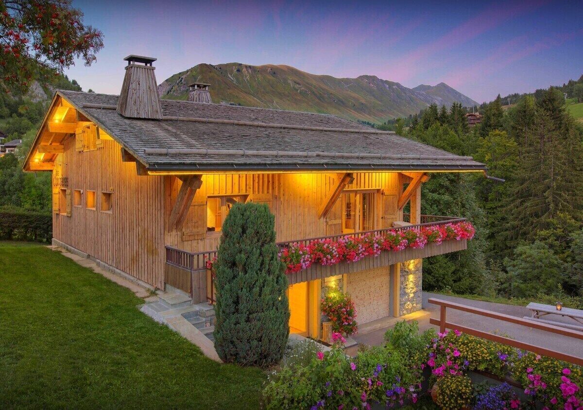 A wooden chalet with a balcony, surrounded by greenery and mountains.