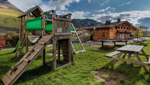 Una struttura giochi in legno con scivolo e rete da arrampicata, un’area picnic con panche e un edificio rustico in legno immerso nella campagna.