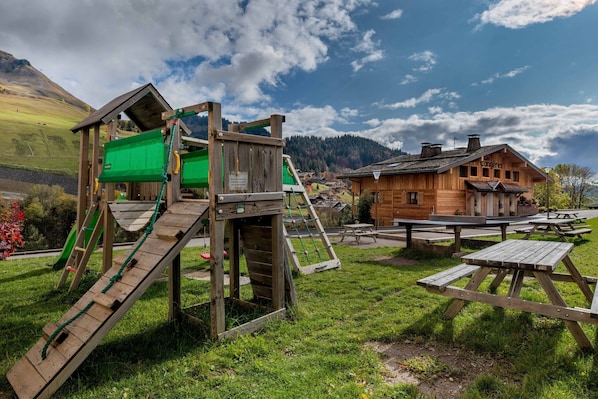 Eine Holzspielanlage mit Rutsche und Kletterbereich, Picknicktische und ein Chalet im Hintergrund.