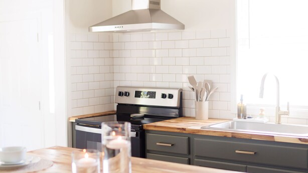 A modern kitchen with white tiled walls, a stainless steel range hood, a wooden dining table set with white cups and saucers, and a window providing natural light. - Cozy Historic District Cottage (Dothan)