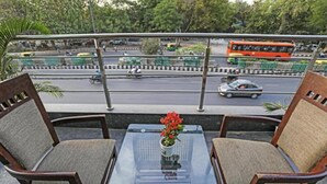Un balcón con dos sillas de madera y una mesa de vidrio, con vista a una calle concurrida con vehículos y vegetación.
