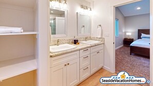 Une salle de bain moderne avec deux lavabos, un meuble-lavabo et un grand miroir. Un dressing se trouve à gauche, et une chambre est visible à travers une porte ouverte.