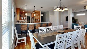 A modern kitchen with a central island, white bar stools, and a dining table with white chairs. - Beaches Be Crazy | Dog Friendly + Game Room & Walk to Beach! (Carolina Beach)