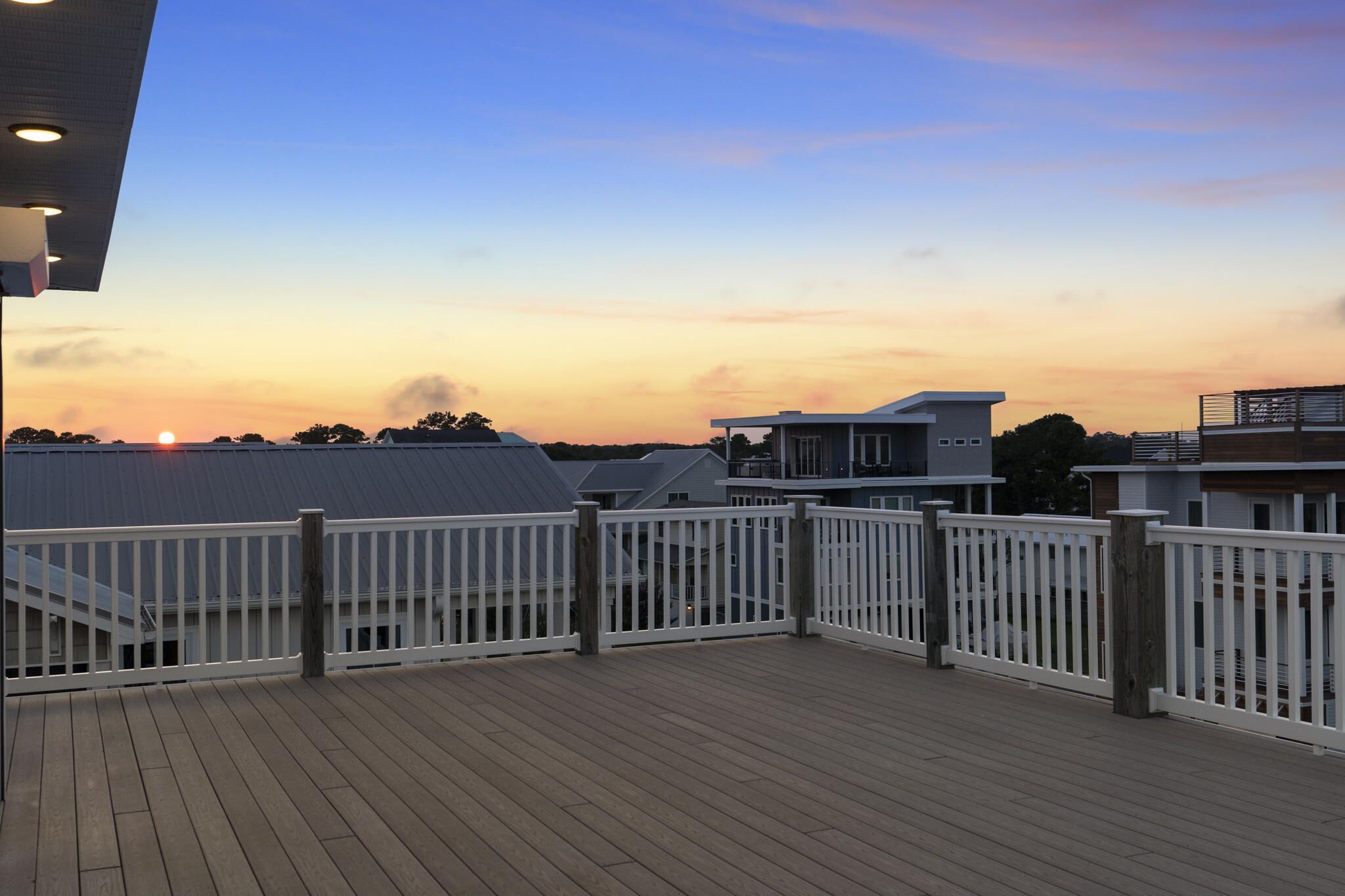 A wooden deck with white railings, overlooking a residential area with houses and a sunset in the background.
