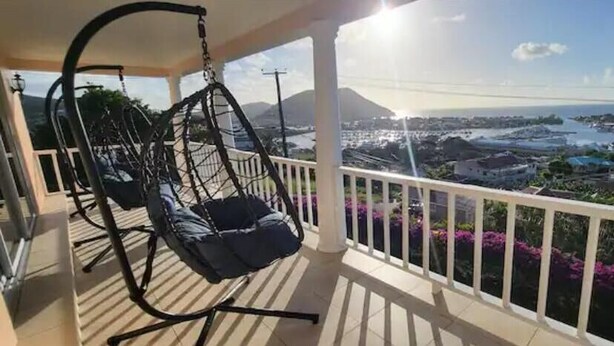 A balcony with a view of a coastal city, a hanging chair, and a railing. - Marina View Villa (Gros Islet)