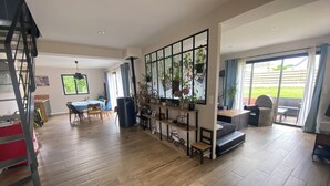 Un'ampia zona giorno con pavimento in legno, un tavolo da pranzo e un angolo salotto con divano e poltrone. Una grande finestra offre luce naturale e vista sull'esterno.