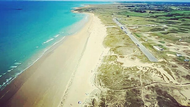 Vista aerea di una zona costiera con spiaggia, una strada e edifici sparsi.