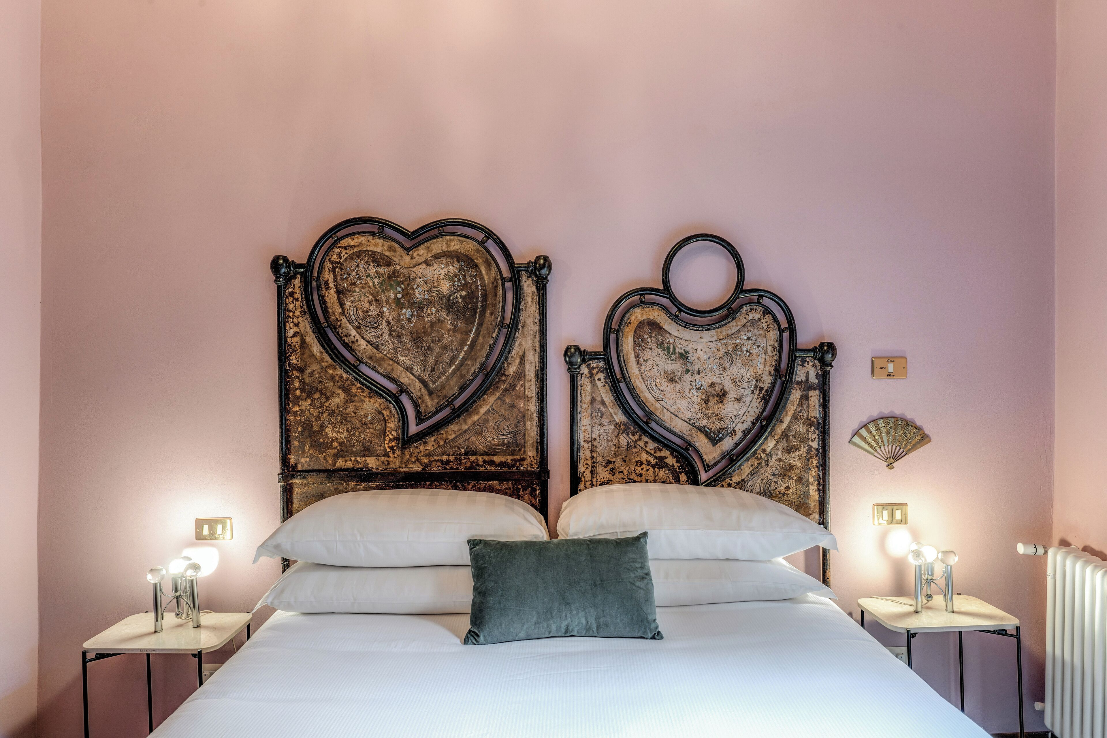 A bedroom with a large bed, two nightstands, a decorative headboard, and a small table with a fan.