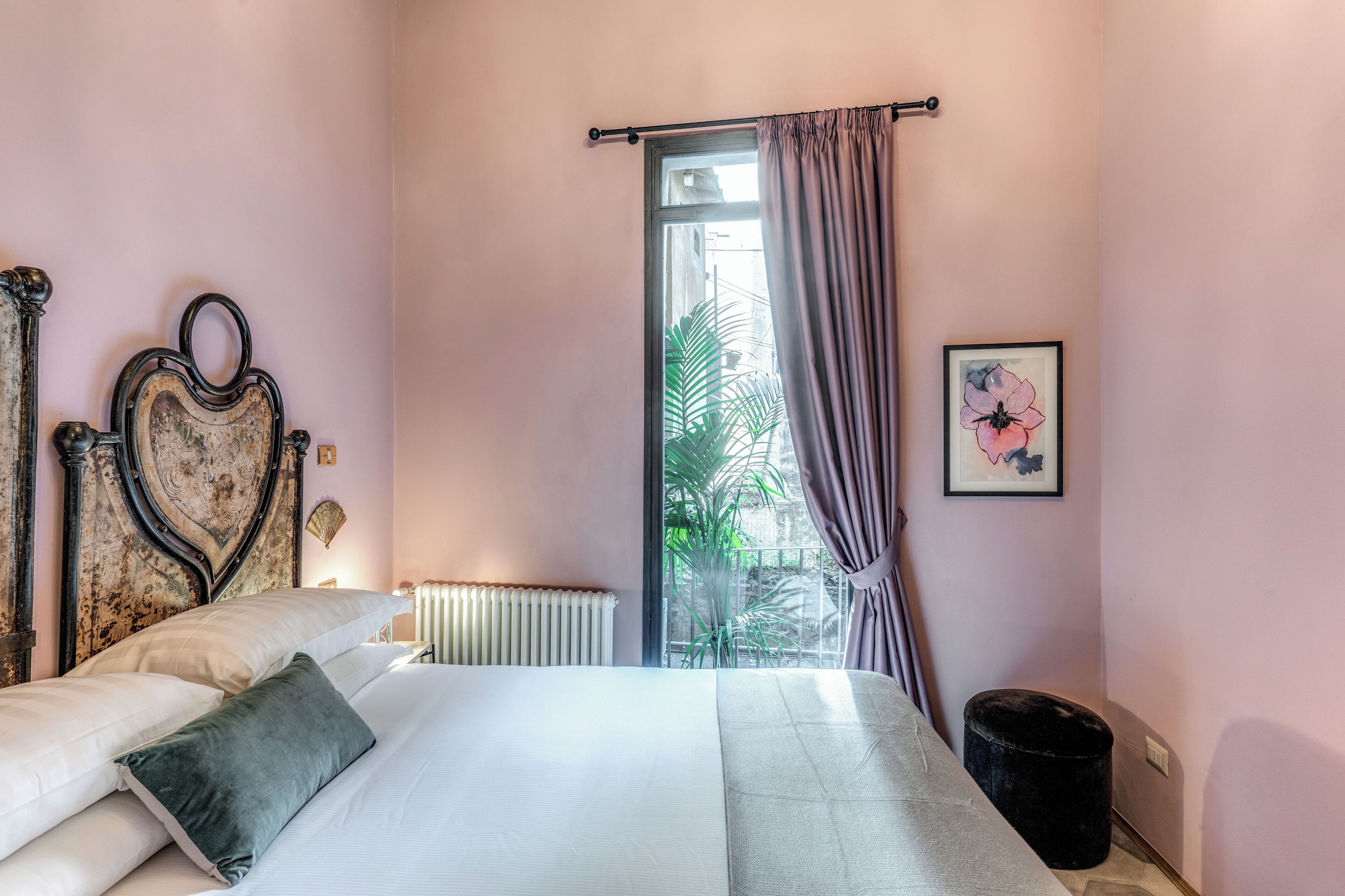 A bedroom with a bed, a radiator, a window with curtains, a framed picture, and a black trash bin.
